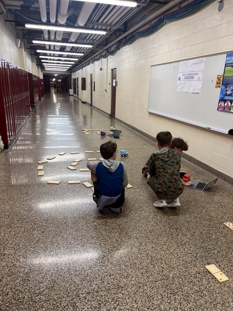 Kids in a hallway watching cars run between dominoes